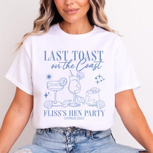 May include: A white short-sleeve t-shirt featuring a blue graphic print of three cocktail glasses, seashells, and stars. The text reads LAST TOAST on the Coast, FLISS'S HEN PARTY, and CYPRUS 2027 in a mix of serif and script fonts.