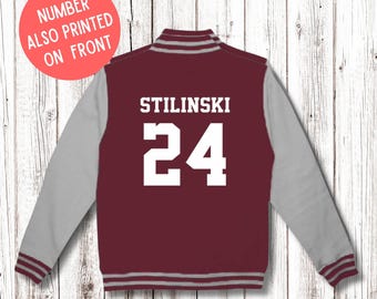 May include: Maroon and gray letterman jacket with the name "STILINSKI" and the number "24" in white block letters on the back. The sleeves are gray, and the jacket has striped trim. A red circle with the text "NUMBER ALSO PRINTED ON FRONT" is in the upper left.