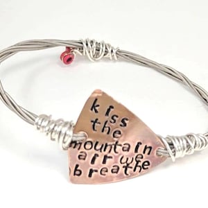 May include: A handmade bracelet crafted from silver-colored guitar strings. The bracelet features a copper-colored triangular charm with the words "kiss the mountain air we breathe" stamped on it. A small red bead accents the design.