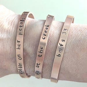 May include: Three copper bracelets with different engraved text. The first bracelet reads "the fall'm on her petals so", the second bracelet reads "meet me at the creek", and the third bracelet reads "b m fs 333".