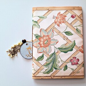 May include: A rectangular notebook with a cream-coloured cover featuring a bamboo lattice pattern, orange and green floral illustrations, and a dangling charm consisting of a small blue circular tag with a palm tree icon and beaded tassels.