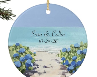 Puede incluir: Adorno redondo de cerámica con una escena de playa. Presenta un cielo azul claro, agua turquesa y un camino arenoso bordeado de flores azules. El texto "Sara & Collin" y la fecha "10-24-26" están impresos.