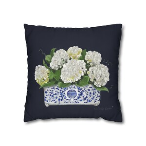 May include: A square throw pillow with a dark navy blue background featuring a central illustration of white hydrangea flowers in a rectangular blue and white patterned ceramic planter with a decorative symbol and the text Girl in her Garden.