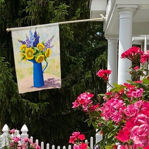 May include: A garden flag displays a painting of sunflowers and blue delphiniums in a blue pitcher. The flag is hanging outside, with a white picket fence and pink roses in the foreground and green trees in the background.