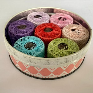 May include: A round tin container with a pink and white chequered pattern holds eight spools of colourful thread. The spools are in shades of purple, pink, red, green, and blue.