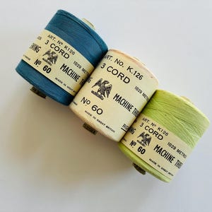 May include: Three spools of vintage machine thread in blue, cream, and green. Each spool has a label with text including "ART. NO. K126", "3 CORD", and "MACHINE THREAD". The spools are labeled "No 60" and "1829 METRES".