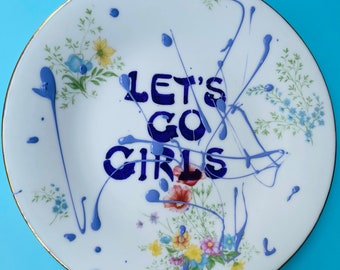 May include: A white ceramic plate with a floral pattern and blue paint splatters. The words "Let's Go Girls" are written in blue paint on the plate.