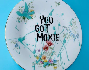 May include: A white porcelain plate with a floral pattern and blue paint splatters. The text "YOU GOT MOXIE" is written in black paint on the plate.