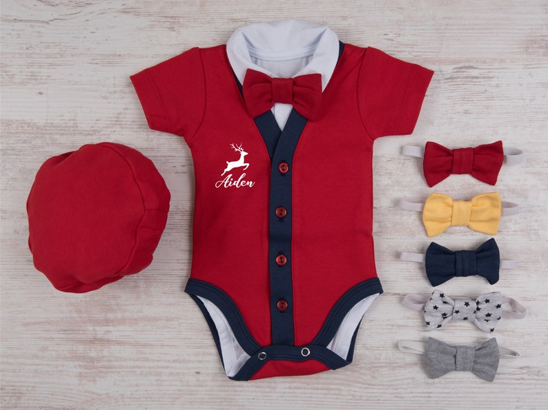 Puede incluir: Un body rojo de manga corta con cuello blanco, pajarita roja y tapeta azul oscuro con cuatro botones rojos. El pecho muestra un gr&aacute;fico de ciervo blanco y el nombre Aiden. Incluye una gorra roja y cinco pajaritas adicionales.