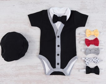 Puede incluir: Un body negro con ribete gris y cuello blanco. El body tiene cierre de botones y una corbata de lazo. Hay cinco corbatas de lazo de diferentes colores y patrones sobre una superficie blanca.