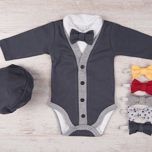May include: A gray bodysuit with a white collar and a gray cardigan. The bodysuit has a button closure and a gray trim. The cardigan has a gray trim and a gray button closure. There are four bow ties in different colors: yellow, red, gray, and gray with white stars. There is also a gray hat.