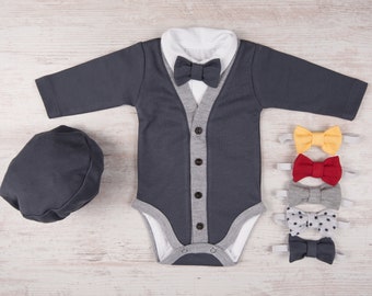 Puede incluir: Un body gris con cuello blanco y un cárdigan gris. El body tiene cierre de botones y un ribete gris. El cardigan tiene un ribete gris y un cierre de botones gris. Hay cuatro pajaritas de diferentes colores: amarillo, rojo, gris y gris con estrellas blancas. También hay un gorro gris.