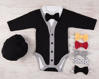 Puede incluir: Un conjunto de bebé negro y gris con cuello blanco y pajarita negra. El conjunto se acompaña de un gorro negro y cuatro pajaritas de diferentes colores y patrones.