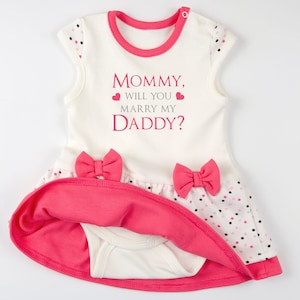 May include: A white sleeveless dress with a pink trim and two pink bows at the waist. The skirt features a white fabric with small black and pink polka dots. The text "MOMMY, WILL YOU MARRY MY DADDY?" is printed in pink and gray on the chest.