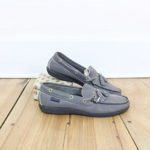 May include: A pair of gray leather boat shoes with tassels. The shoes have a rubber sole and a leather upper. The brand name "DUCKS" is visible on the side of the shoe.
