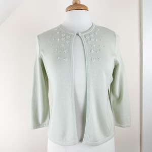 May include: A light green cardigan with a delicate floral embroidery design around the neckline. The cardigan has a long open front and three-quarter sleeves.