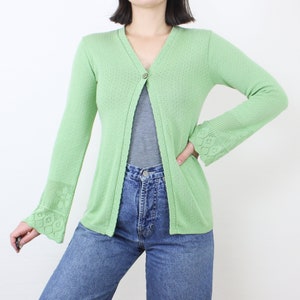 May include: A light green knitted cardigan with a single button closure and bell sleeves. The cardigan has a delicate openwork pattern.
