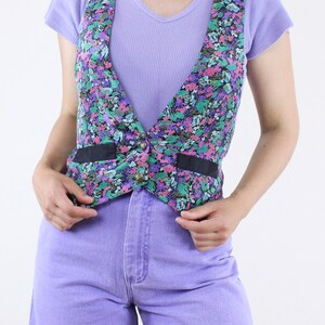 May include: A floral print vest with black ribbon trim and a single button closure. The vest is worn over a lavender tank top and paired with lavender denim shorts.