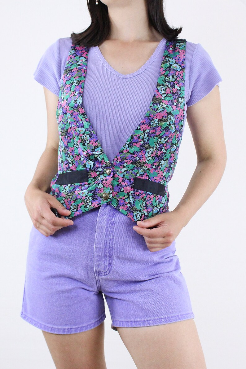 May include: A floral print vest with black ribbon trim and a single button closure. The vest is worn over a lavender tank top and paired with lavender denim shorts.