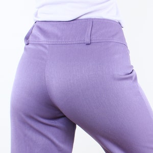 May include: Close-up of lavender-colored pants with a high waist and belt loops. The pants are made of a smooth fabric and have a tailored fit. The pants are part of a clothing collection.