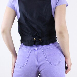 May include: A black vest with a satin finish and a button closure. The vest is worn over a light purple shirt and paired with lavender denim shorts.