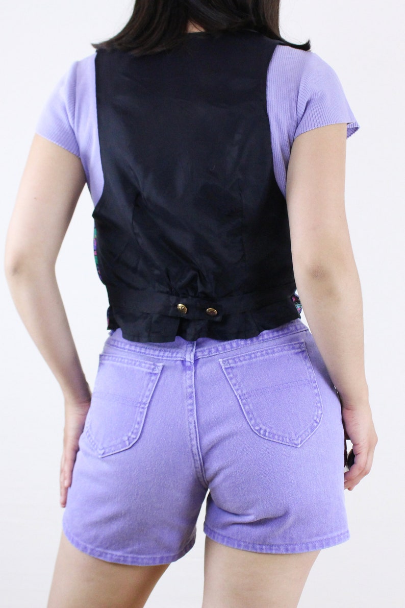 May include: A black vest with a satin finish and a button closure. The vest is worn over a light purple shirt and paired with lavender denim shorts.