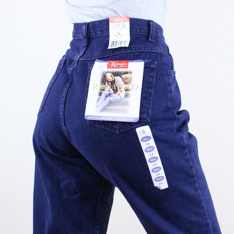 Może przedstawiać: Jeans z ciemnoniebieskiego denimu z przyczepioną do tylnej kieszeni metką Wrangler Relaxed Fit. Na metce widnieje kobieta w tych dżinsach i napis "Relaxed Fit".