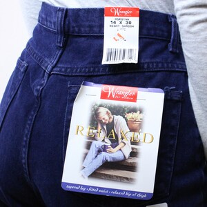 Może przedstawiać: Dżinsy w ciemnym odcieniu niebieskiego z metką Wrangler. Na metce widnieje napis "Relaxed" oraz "tapered leg, fitted waist, relaxed hip & thigh". Dżinsy mają metkę z rozmiarem "14 x 30" i "Night Shadow Short".