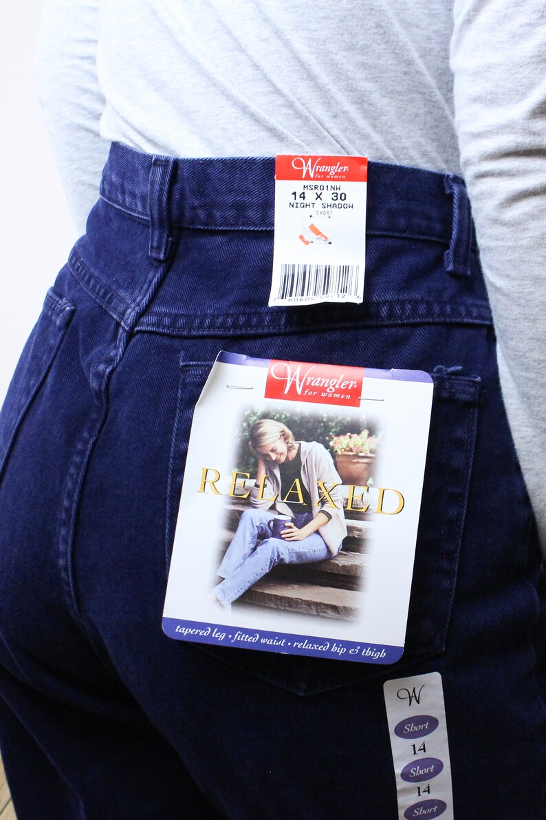Może przedstawiać: Dżinsy w ciemnym odcieniu niebieskiego z metką Wrangler. Na metce widnieje napis "Relaxed" oraz "tapered leg, fitted waist, relaxed hip & thigh". Dżinsy mają metkę z rozmiarem "14 x 30" i "Night Shadow Short".