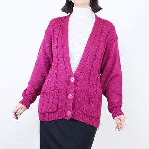 May include: A magenta knitted cardigan with a cable knit pattern, long sleeves, and a v-neck. The cardigan has four buttons down the front and two pockets.