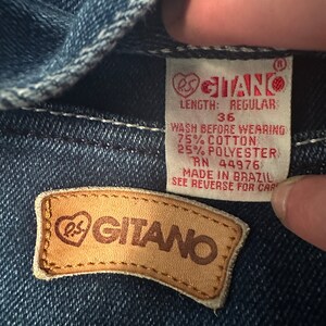 May include: A close-up of a blue denim jean label with the text "RS GITANO" in a heart-shaped logo. The label also includes the text "LENGTH: REGULAR", "36", "WASH BEFORE WEARING", "75% COTTON", "25% POLYESTER", "RN 44976", "MADE IN BRAZIL", and "SEE REVERSE FOR CARE".