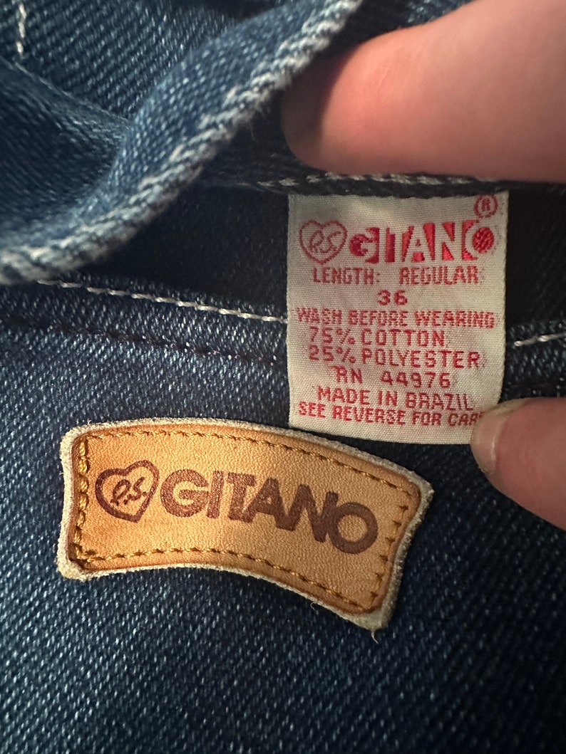 May include: A close-up of a blue denim jean label with the text "RS GITANO" in a heart-shaped logo. The label also includes the text "LENGTH: REGULAR", "36", "WASH BEFORE WEARING", "75% COTTON", "25% POLYESTER", "RN 44976", "MADE IN BRAZIL", and "SEE REVERSE FOR CARE".