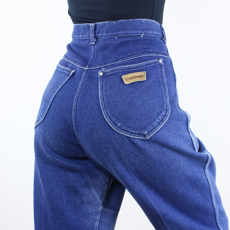 May include: A pair of high-waisted blue denim jeans with a back pocket featuring a brown leather patch with the word "GITANO" in white lettering.