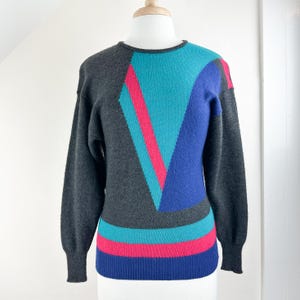 May include: A gray sweater with a geometric pattern in teal, blue, and pink. The sweater has long sleeves and a crew neck.