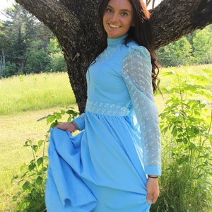 May include: A woman wearing a light blue dress with lace sleeves and a white lace trim. The dress has a ruffled skirt and is worn with a high neckline. The woman is standing in a field with green grass and trees in the background.