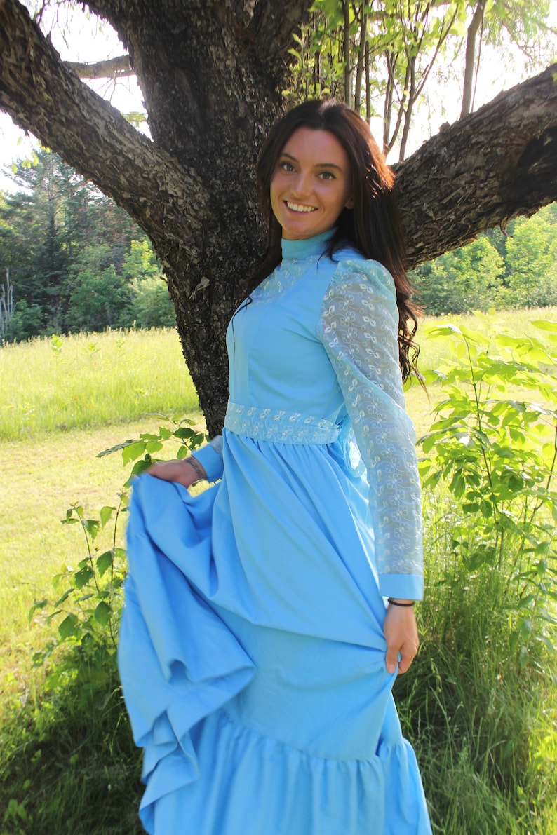 May include: A woman wearing a light blue dress with lace sleeves and a white lace trim. The dress has a ruffled skirt and is worn with a high neckline. The woman is standing in a field with green grass and trees in the background.