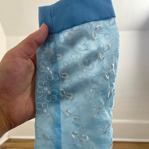May include: Light blue fabric with a white floral pattern and a blue trim. The fabric is sheer and has a delicate texture.