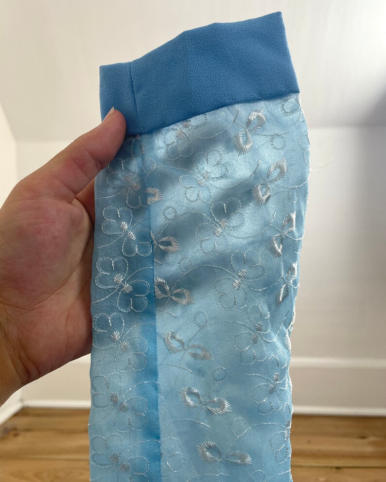 May include: Light blue fabric with a white floral pattern and a blue trim. The fabric is sheer and has a delicate texture.
