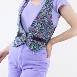 May include: A floral print vest with black trim and a button closure. The vest is worn over a light purple t-shirt and paired with lavender denim shorts.