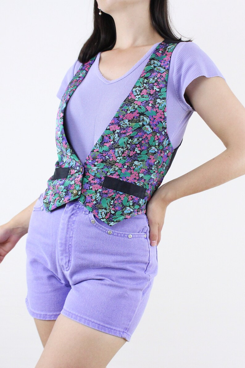 May include: A floral print vest with black trim and a button closure. The vest is worn over a light purple t-shirt and paired with lavender denim shorts.