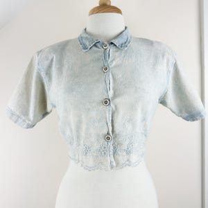 May include: A light blue denim button-up shirt with short sleeves and a white embroidered trim around the bottom.