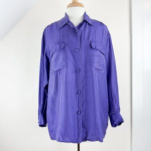 May include: A long-sleeved, purple button-down shirt with two front pockets. The shirt is displayed on a mannequin. The fabric has a slight sheen, suggesting a silk or satin material. The shirt has a classic collar and buttoned cuffs.