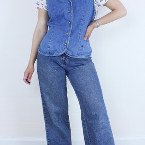 May include: A blue denim vest with a button-up front and a heart-shaped design on the front. The vest is worn over a white floral print shirt and blue jeans.