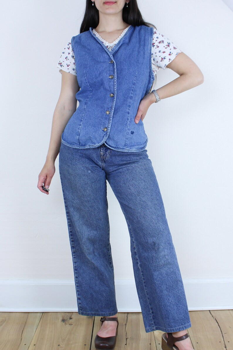 May include: A blue denim vest with a button-up front and a heart-shaped design on the front. The vest is worn over a white floral print shirt and blue jeans.