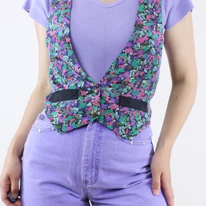 May include: A floral print vest with a black trim and a single button closure. The vest is worn over a lavender ribbed tank top and paired with lavender denim shorts.