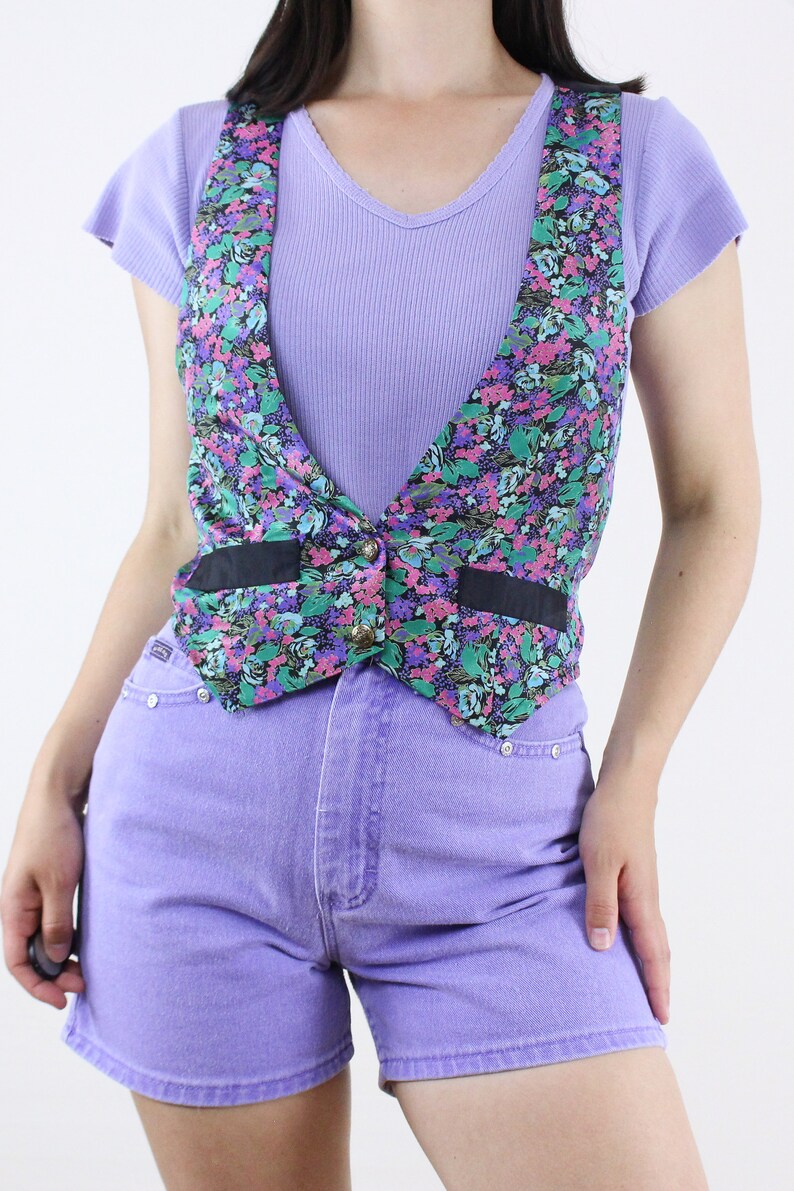 May include: A floral print vest with a black trim and a single button closure. The vest is worn over a lavender ribbed tank top and paired with lavender denim shorts.