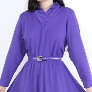 May include: A long-sleeved, purple dress with a V-neckline and a matching belt. The dress has a flowing skirt and a fitted waist, creating a classic silhouette. The belt is a thin, shiny purple with a gold-colored buckle.