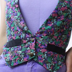 May include: A floral print vest with black trim and gold buttons. The vest is worn over a purple ribbed tank top and purple jeans.