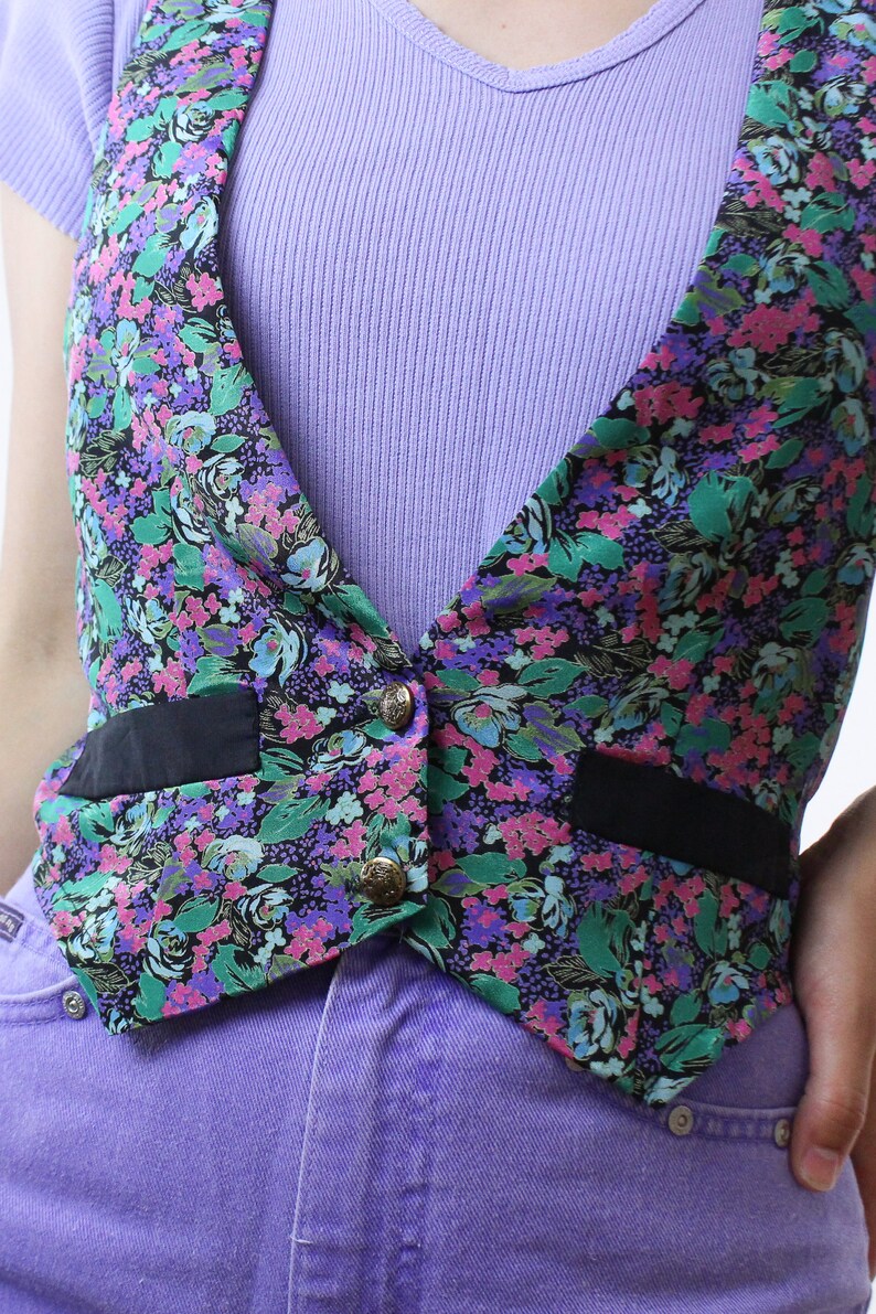May include: A floral print vest with black trim and gold buttons. The vest is worn over a purple ribbed tank top and purple jeans.