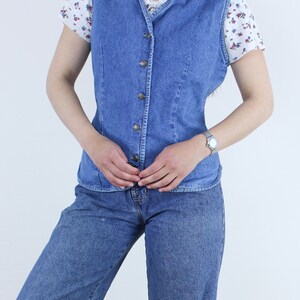 May include: A blue denim vest with a button-up front and a V-neck. The vest is worn over a white floral print shirt and blue jeans.